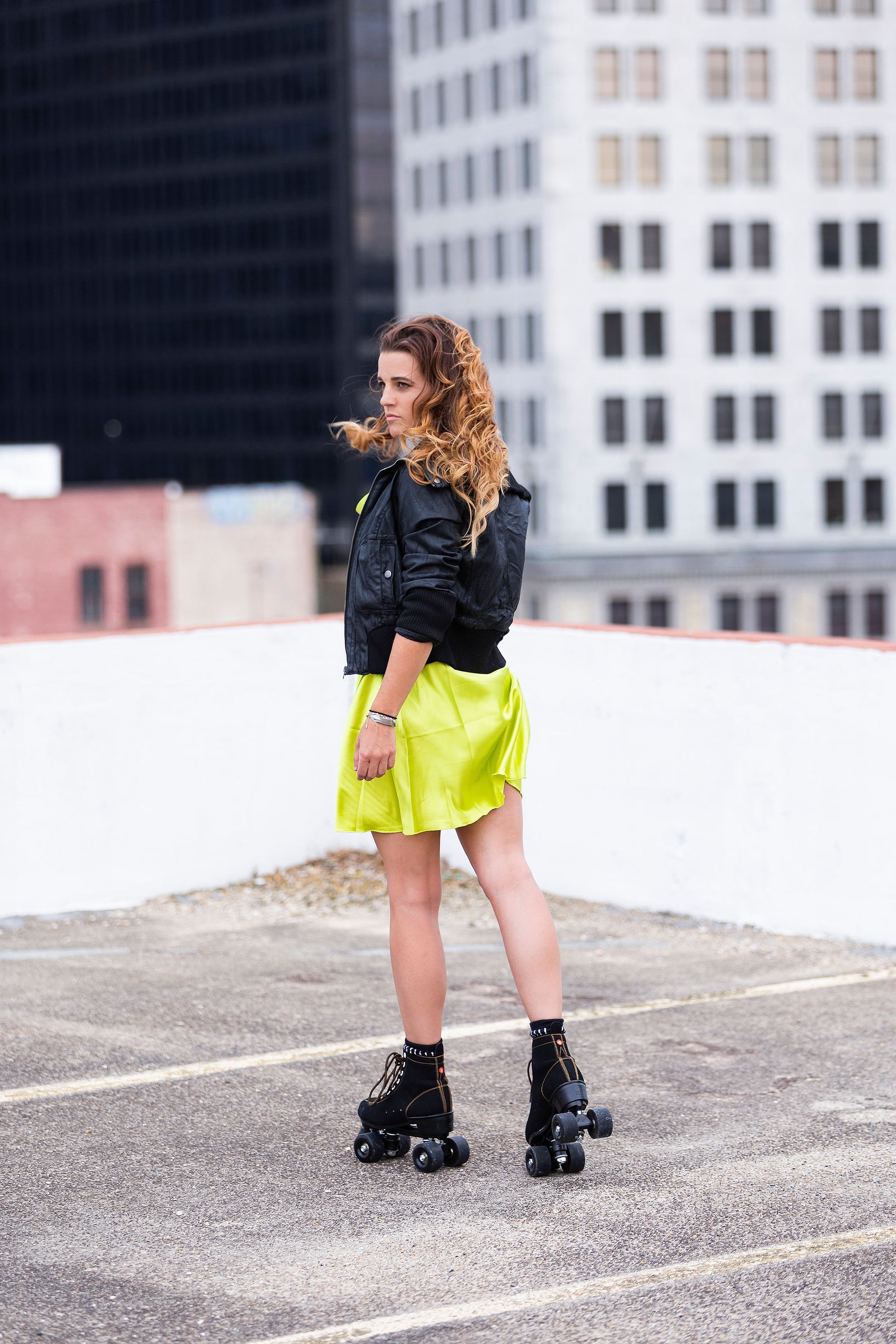 Person in a neon yellow skirt and black roller skates standing on a rooftop with a city skyline in the background.