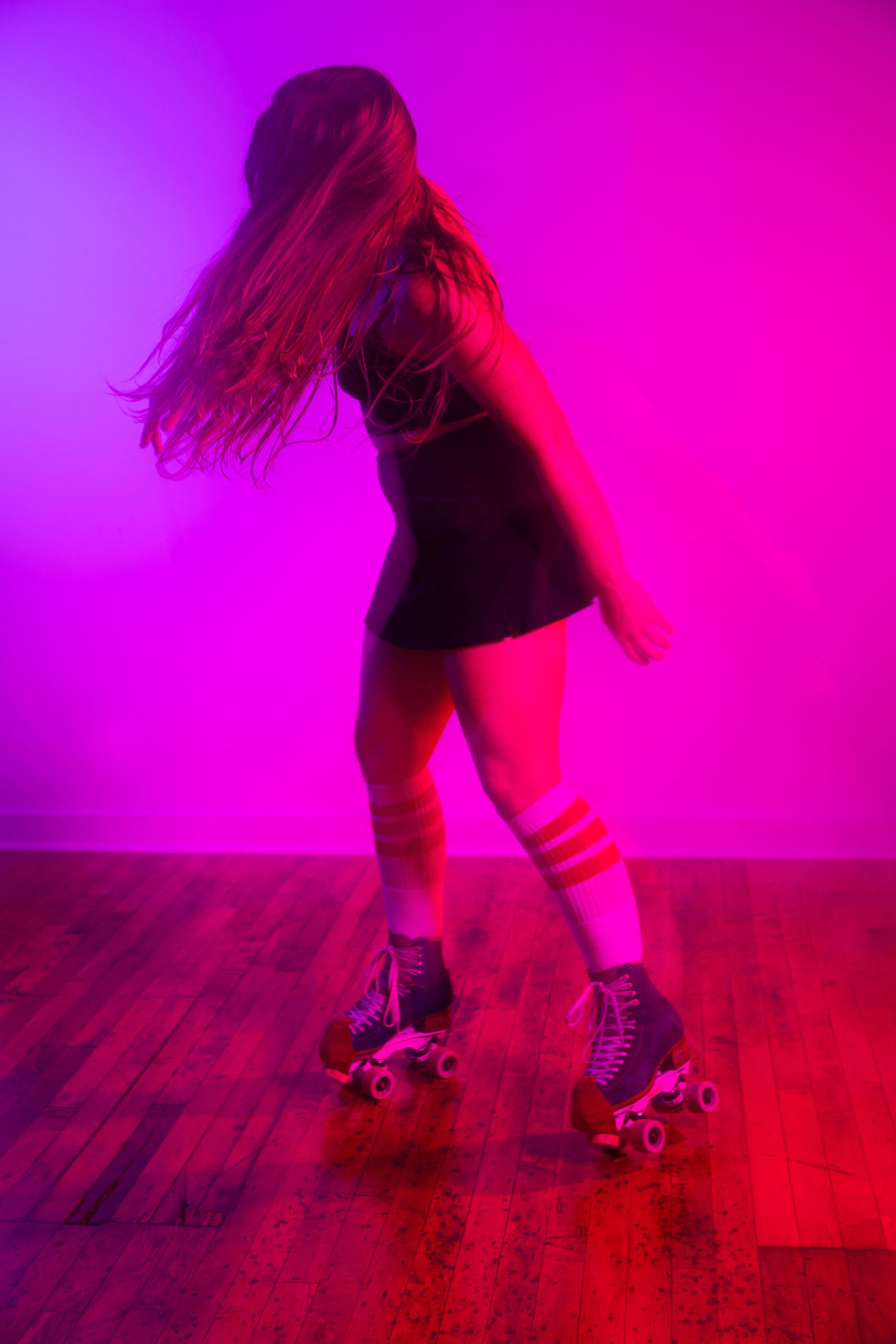 Person in motion on roller skates with long hair flowing, captured under dramatic pink and purple lighting on a wood floor.