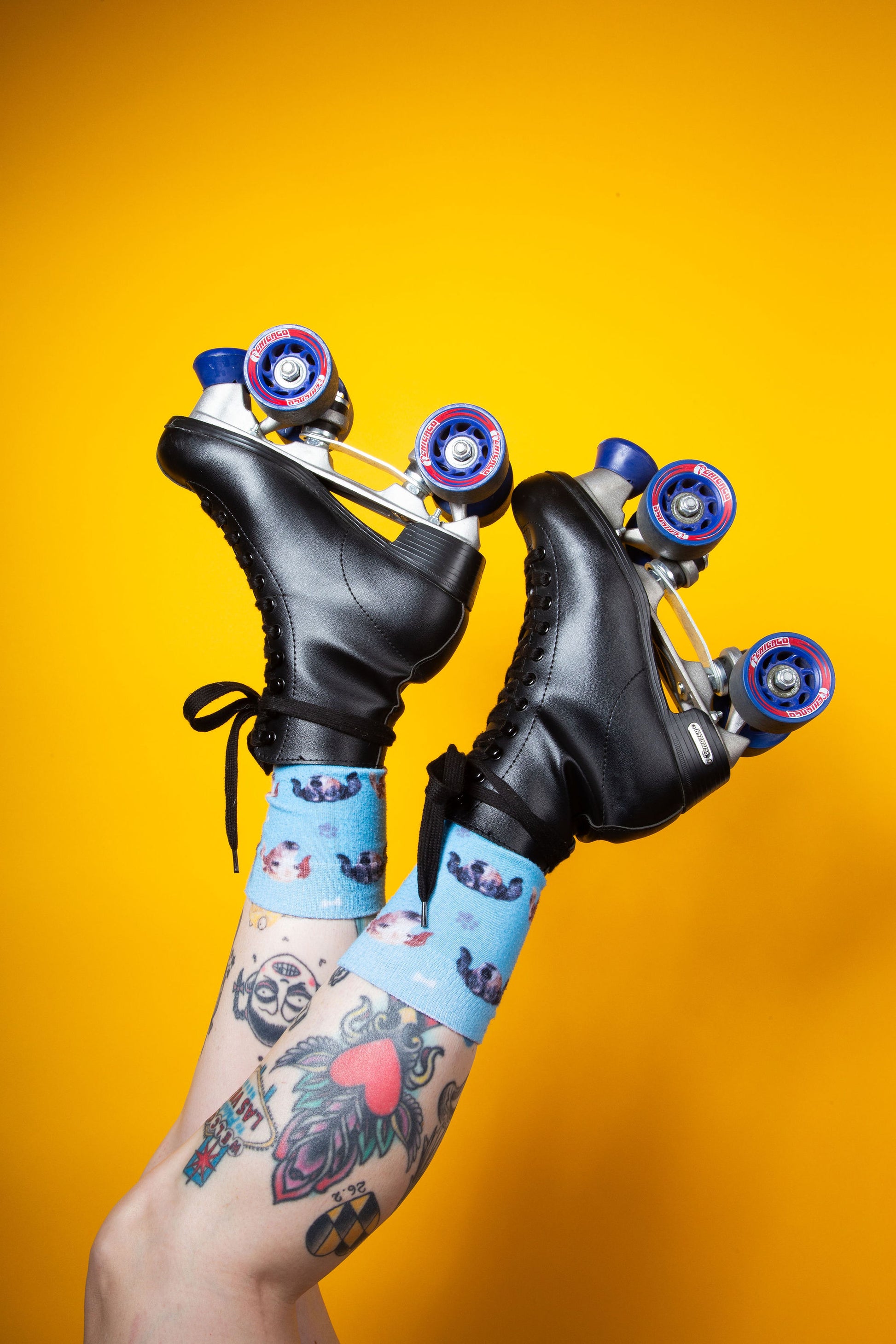Tattooed legs raised in the air wearing black roller skates and blue animal-print socks against a yellow background.