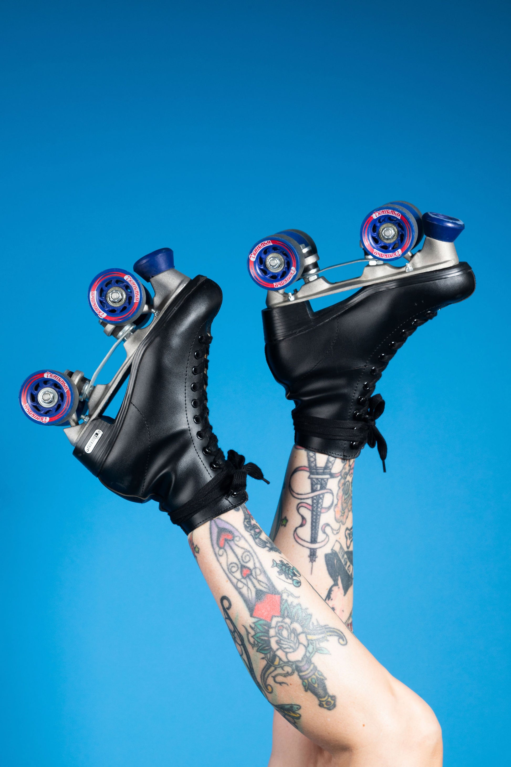 Tattooed legs raised in the air wearing black roller skates set against a blue background.