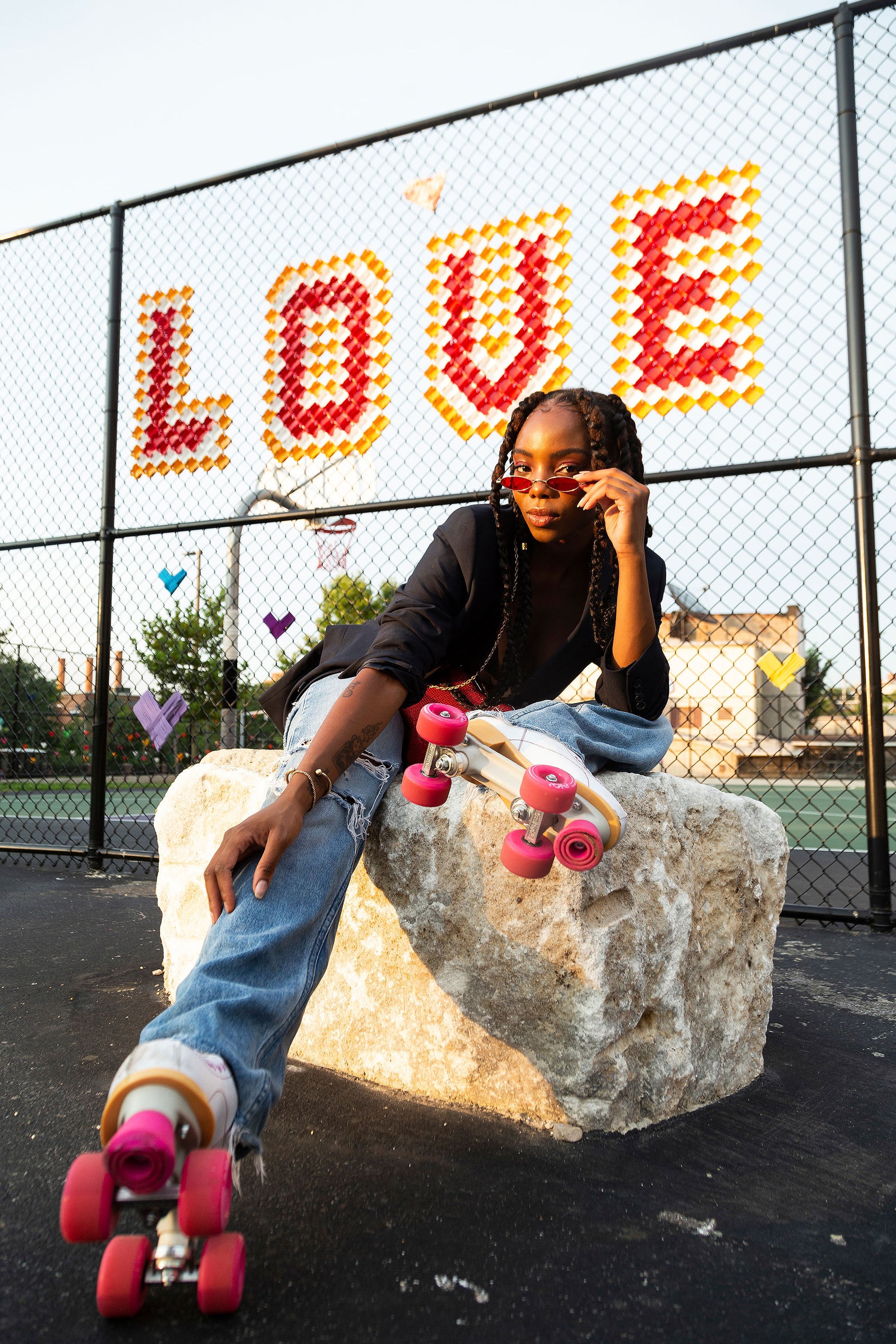 Roller skater lounging on a boulder in front of a chain-link fence spelling “LOVE” in bold red and yellow cups, with vibrant pink skates and a confident gaze.