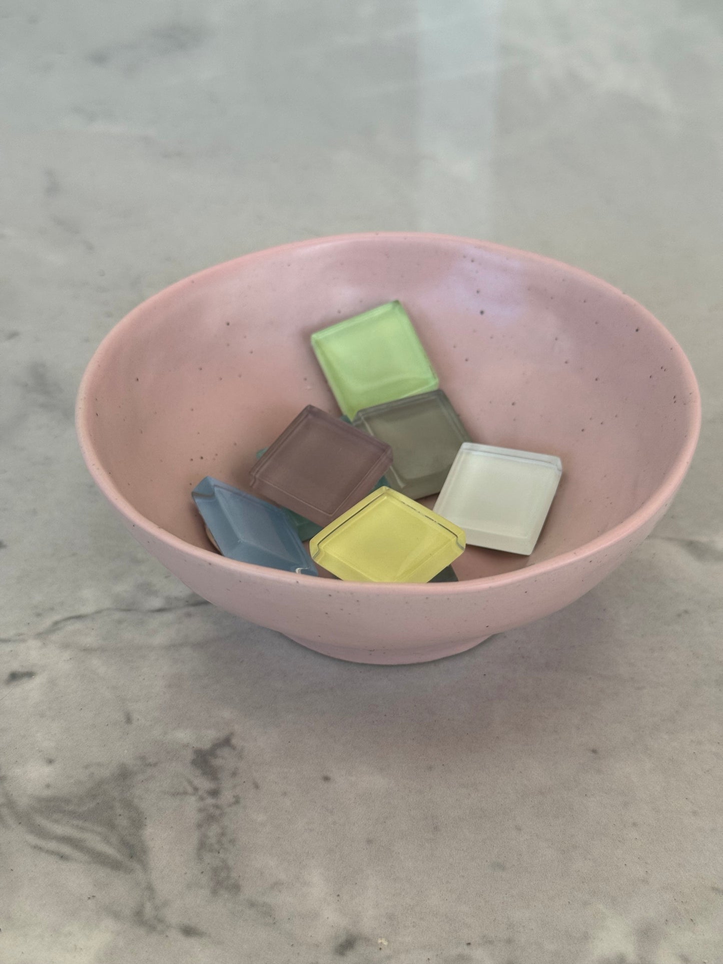 A pink speckled ceramic bowl containing several small square wax melts in assorted pastel and neutral colors, set on a light gray marble surface.