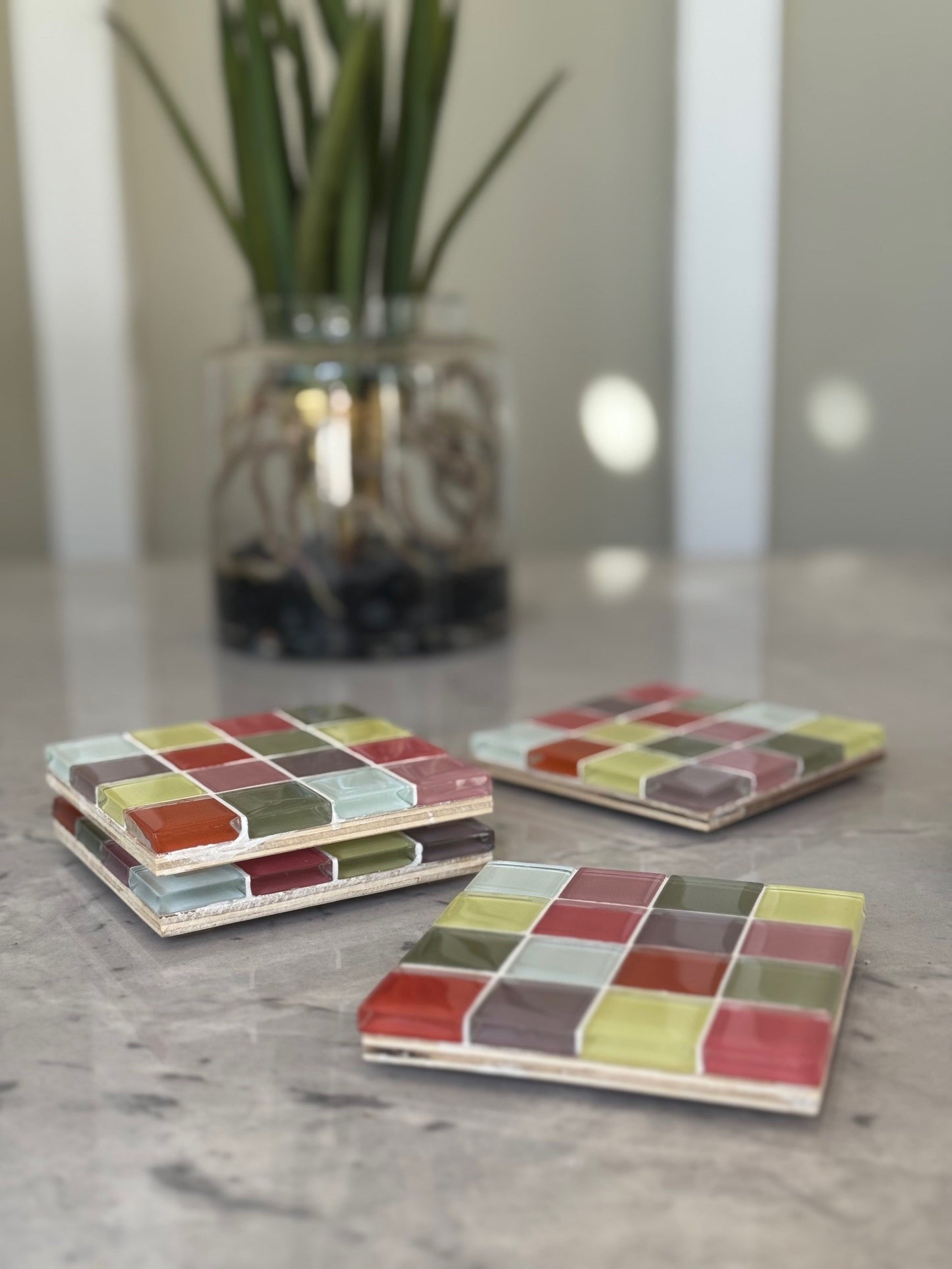 Three square ceramic coasters with a colorful mosaic tile pattern in red, green, yellow, and brown, arranged on a gray marble countertop with a blurred potted plant in the background.