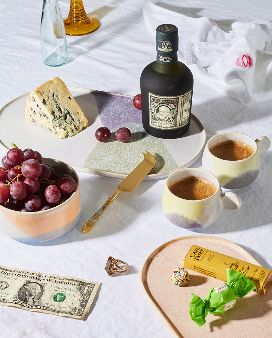 Bottle of Chivas Regal whiskey with a plate of cheese, grapes, and two cups of coffee on a white surface.