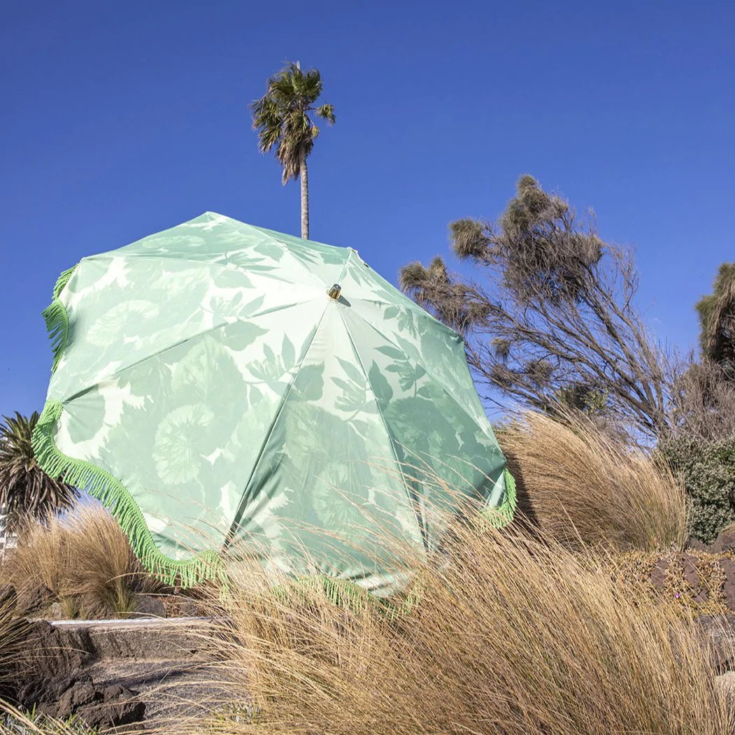 retro inspired beach umbrella in green with floral pattern and green fringes and wooden pole in the dunes with a clear blue sky and a palm tree