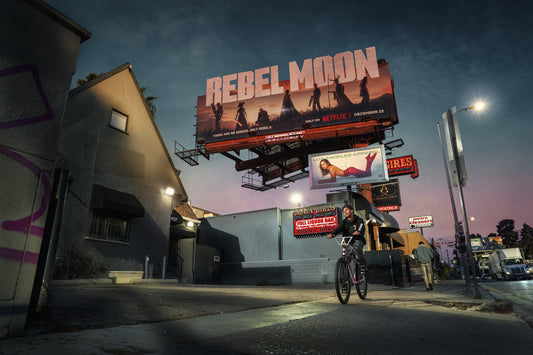 Man on a bicycle in front of a large 'Rebel Moon' billboard at night.