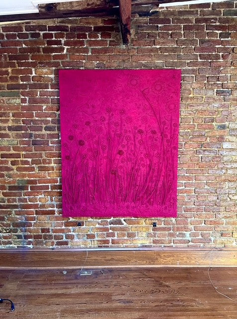 Pink mixed media art work with a floral pattern hanging on a brick wall.