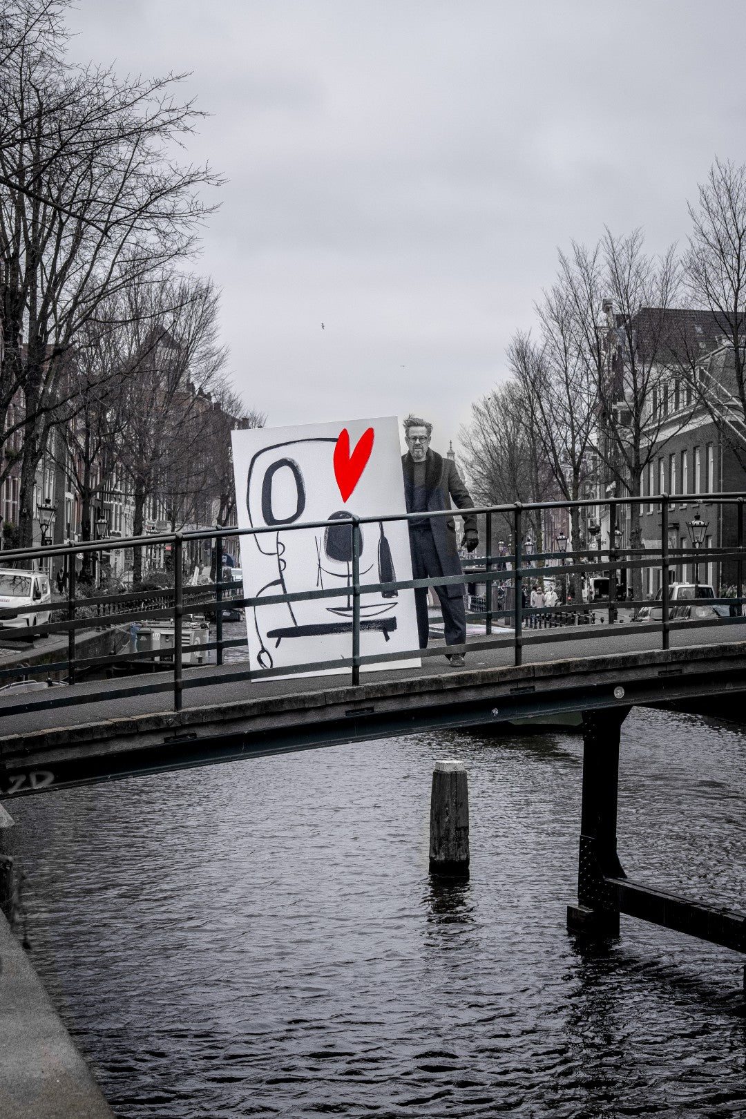 Artist Selwyn Senatori holding a large sign with a red heart over a canal bridge in a Amsterdam