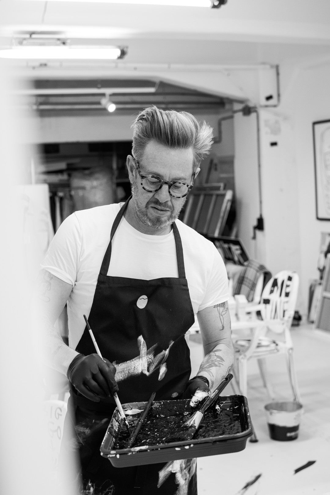 Person wearing an apron and gloves, holding a tray with paintbrushes in a studio setting.