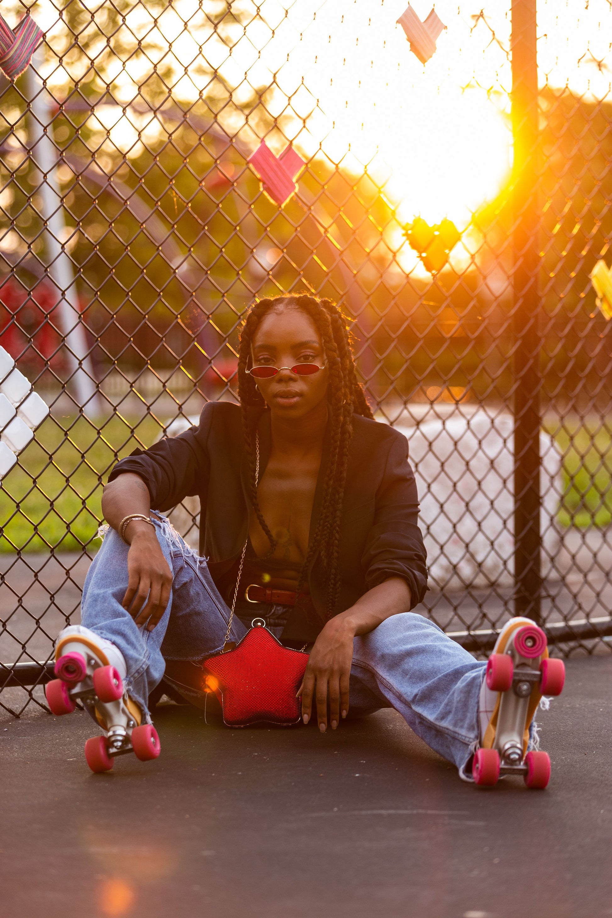 Person in roller skates sitting against a chain-link fence at sunset, wearing sunglasses, a blazer, and ripped jeans with a red star-shaped bag.
