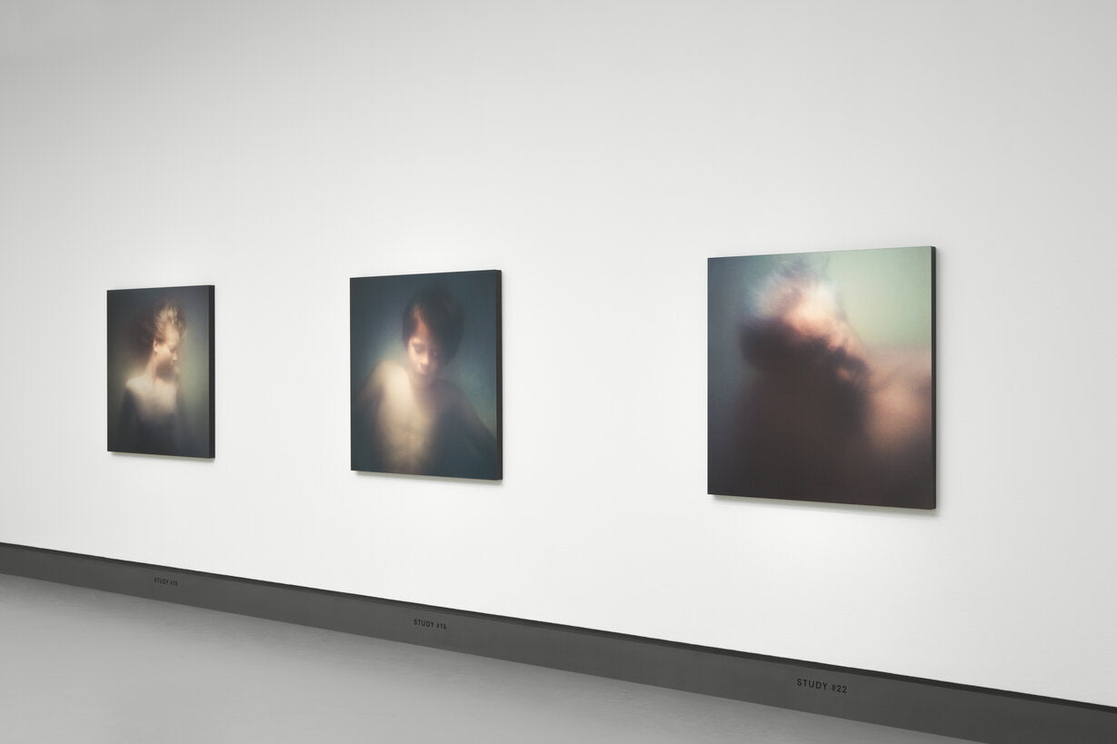 Three framed abstract portraits hang in a gallery setting on a white wall above a dark floor. The artworks feature soft, blurred figures in muted tones of gray, blue, brown, and cream.