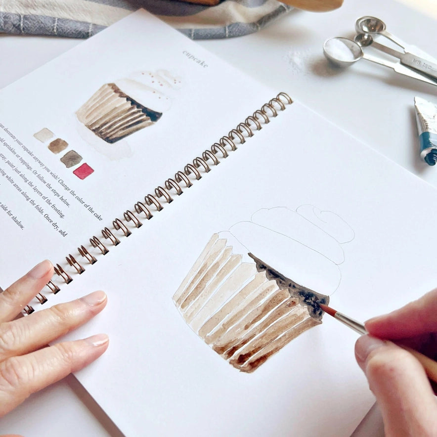 hands holding a brush and water coloring an illustration of a cupcake