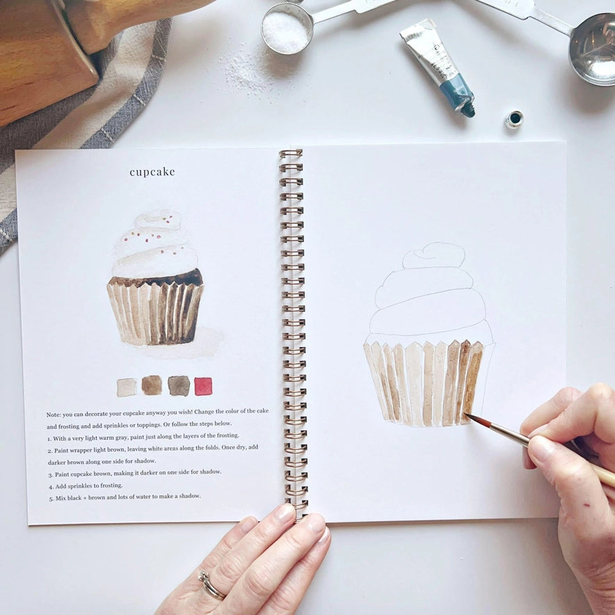 hands holding a brush and water coloring an illustration of a cupcake