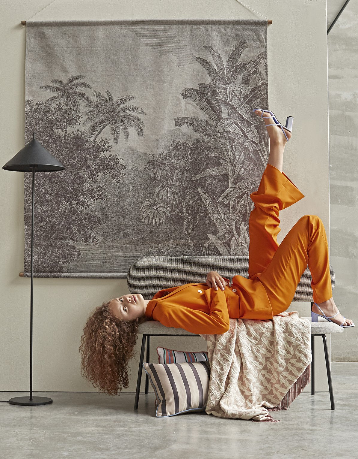 black metal triangle shape floor lamp with wall chart and woman in orange suit on a grey bench