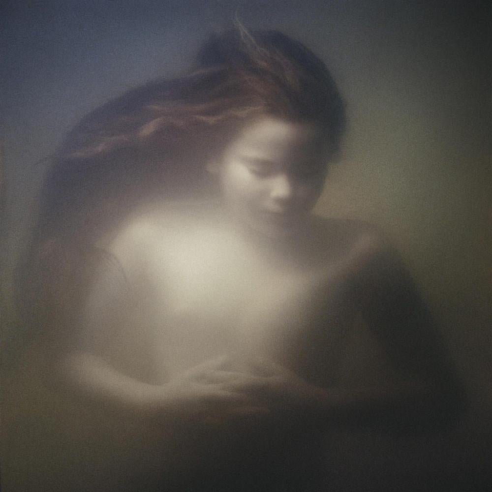 A moody, soft-focus portrait of a woman with long dark hair, rendered in muted gray, brown, and beige tones with a hazy, dreamlike effect.