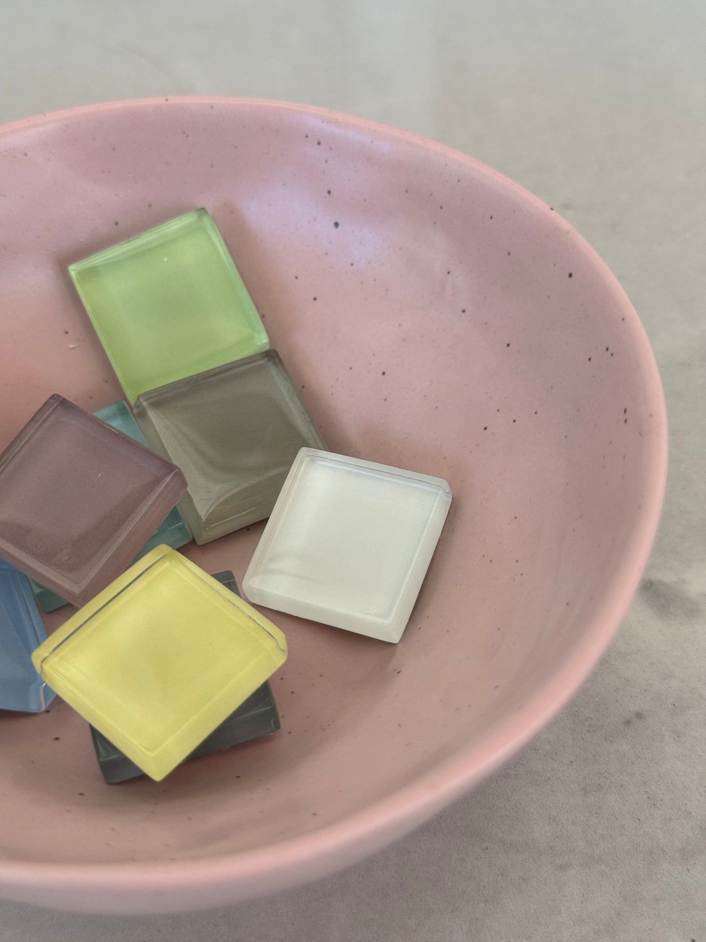 Several small square translucent tiles in pastel colors, including yellow, white, green, brown, and blue, arranged inside a pink speckled bowl on a light surface.