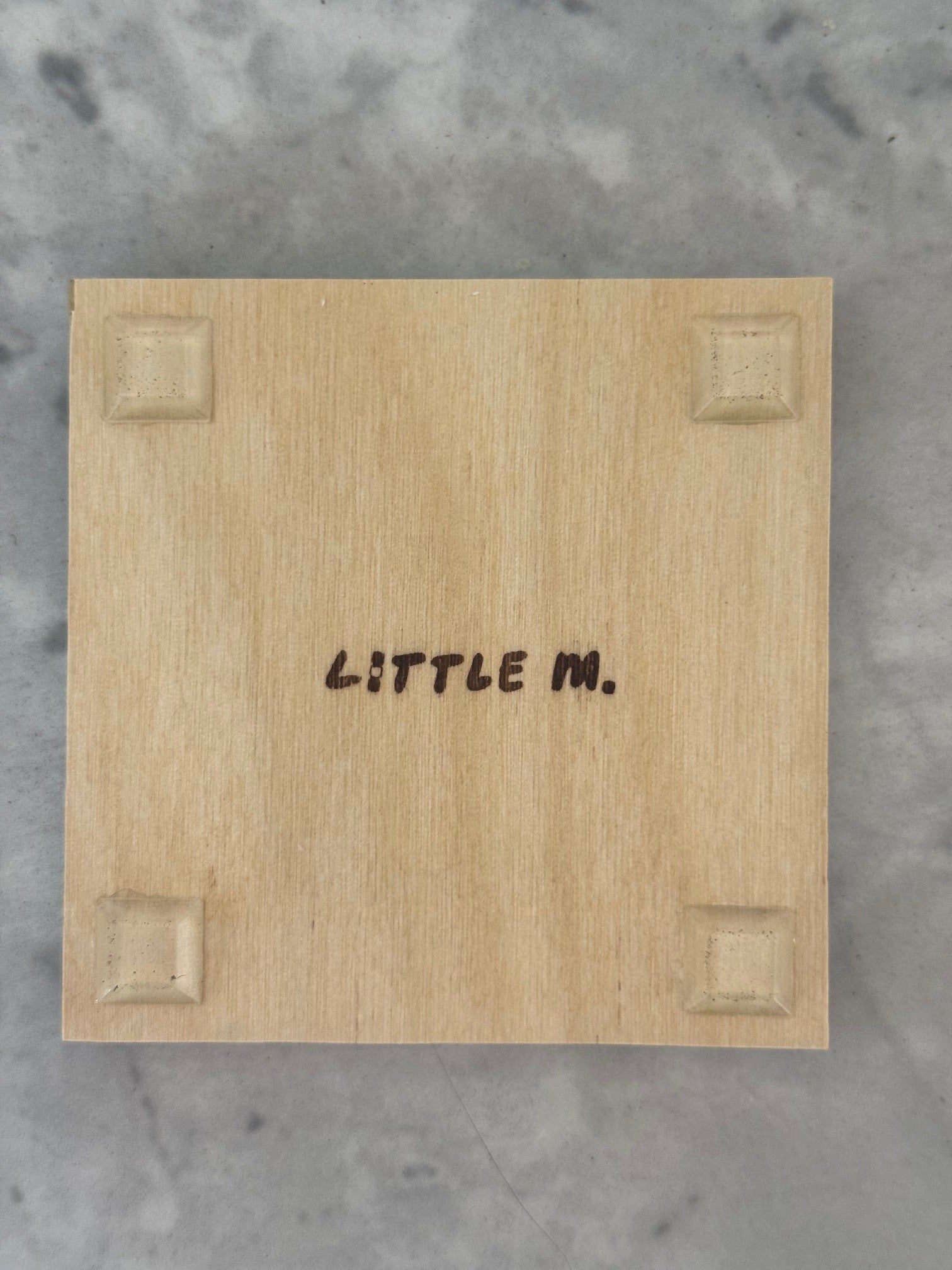 A square light wood block or plaque with the words 'LITTLE M.' printed in black at the center, photographed from above on a gray marble-like surface.