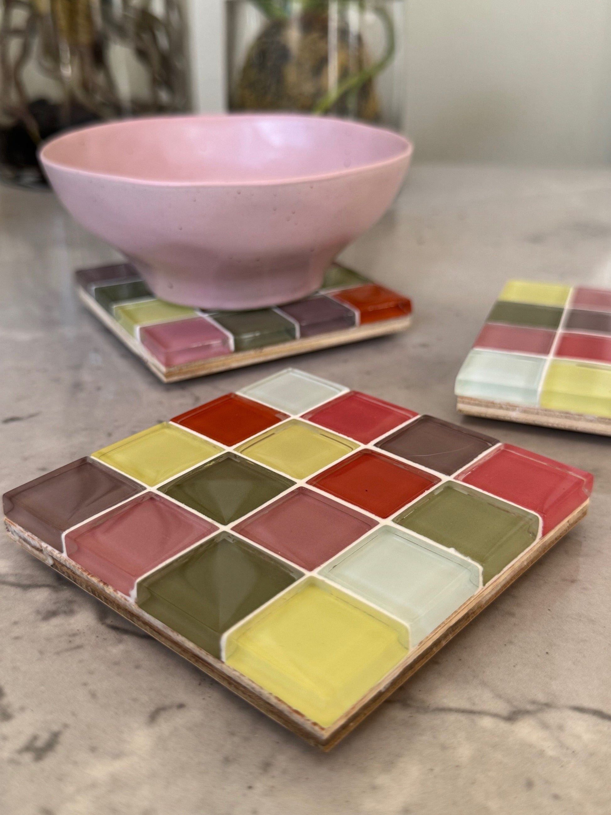A set of square ceramic coasters with a colorful checkered tile pattern in red, yellow, green, and pink, shown on a marble countertop with a pink bowl on one coaster.