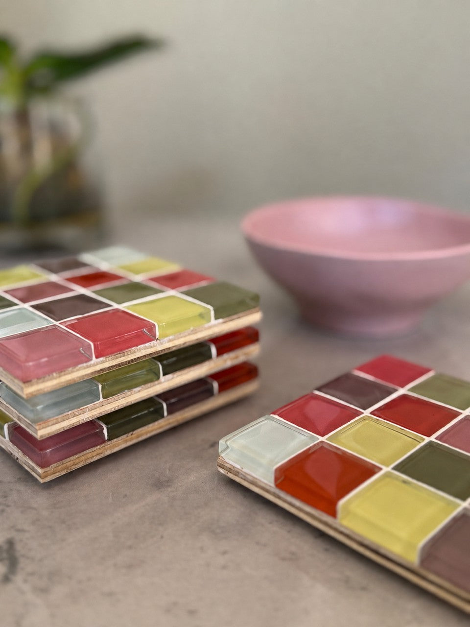 Two square wooden boards with a colorful mosaic tile pattern are arranged on a light gray surface, with a pink bowl and a blurred plant in the background.