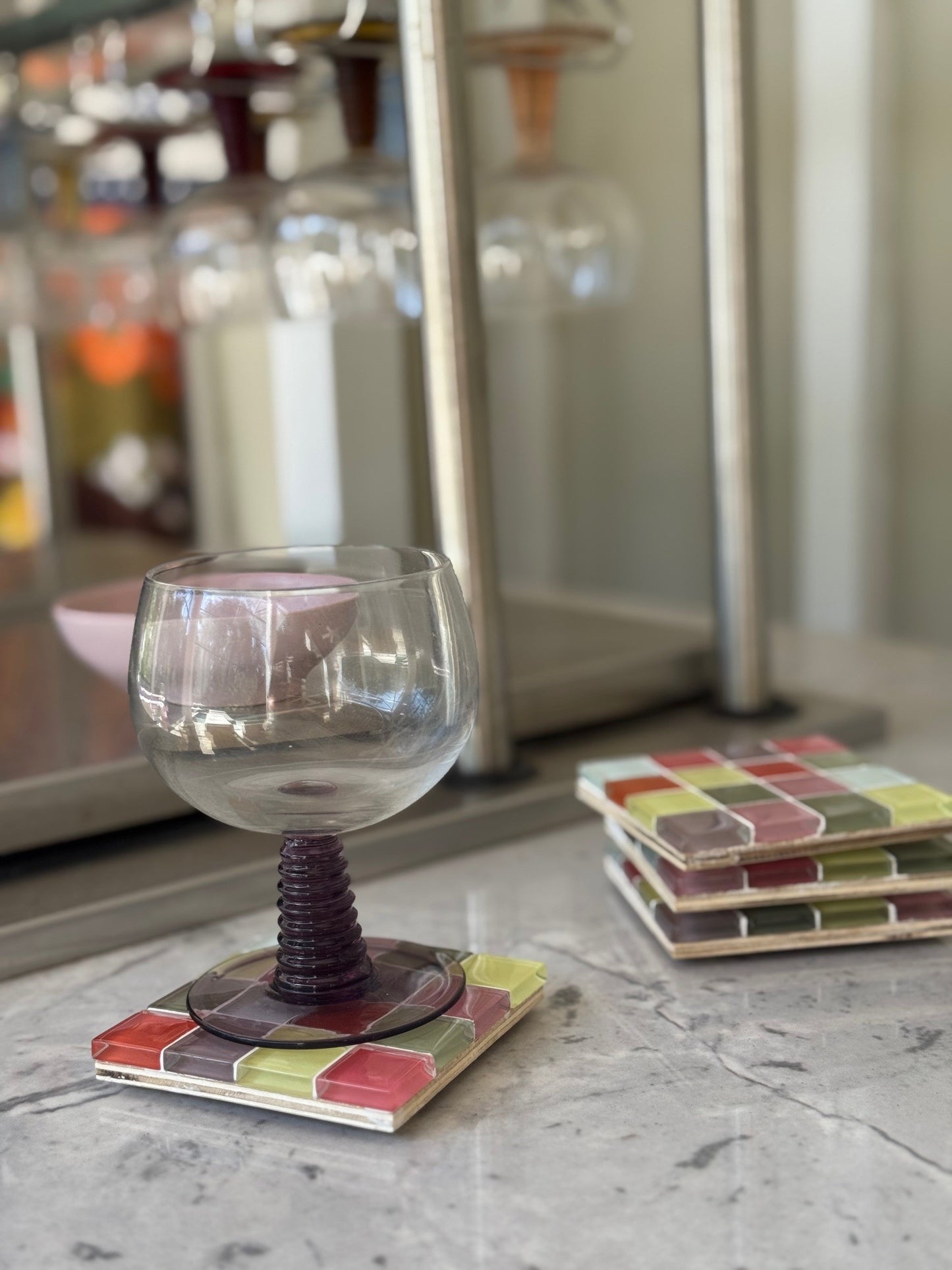 A clear stemmed glass with a dark purple spiral stem sits on a square mosaic coaster, with matching coasters stacked on a marble surface in a softly blurred indoor setting.