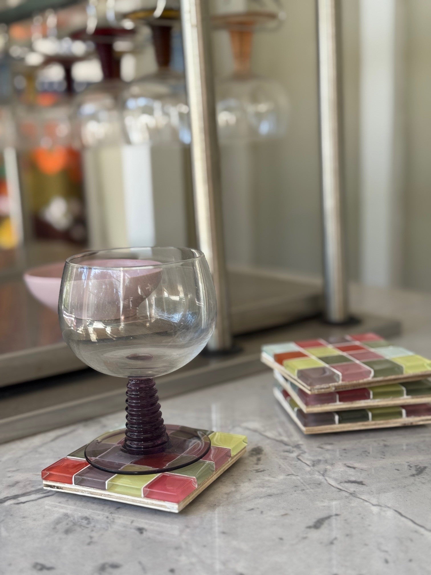 A clear stemmed glass with a dark purple spiral stem sits on a square mosaic coaster, with matching coasters stacked on a marble surface in a softly blurred indoor setting.