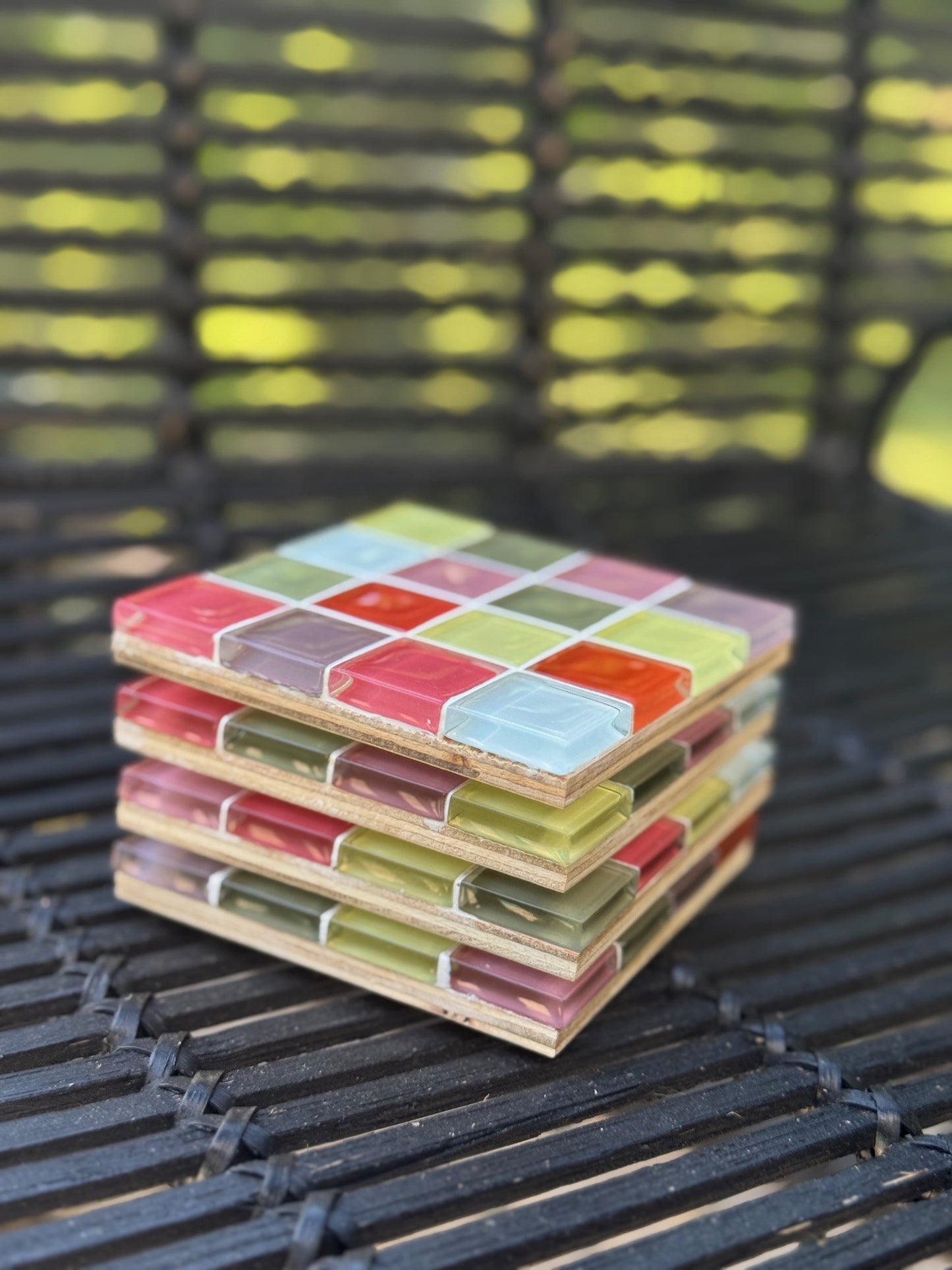 A stack of square wooden coasters with a colorful mosaic tile pattern, resting on a black metal outdoor chair with a blurred green garden background.