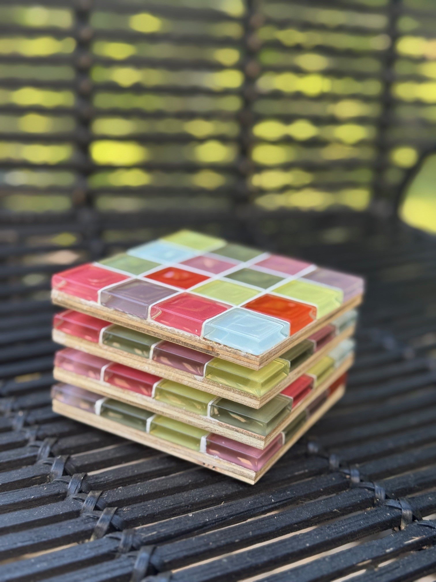 A stack of square wooden coasters with a colorful mosaic tile pattern, resting on a black metal outdoor chair with a blurred green garden background.