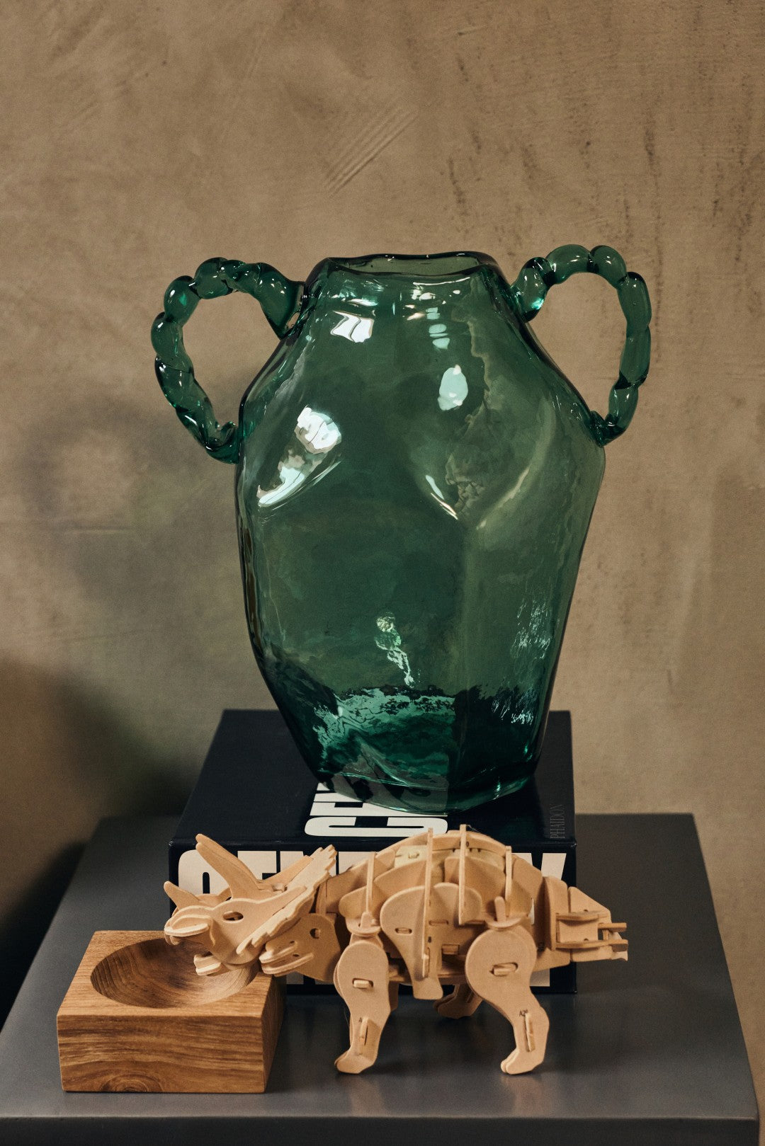 Green glass vase with handles on a stand, accompanied by wooden sculptures on a dark surface.