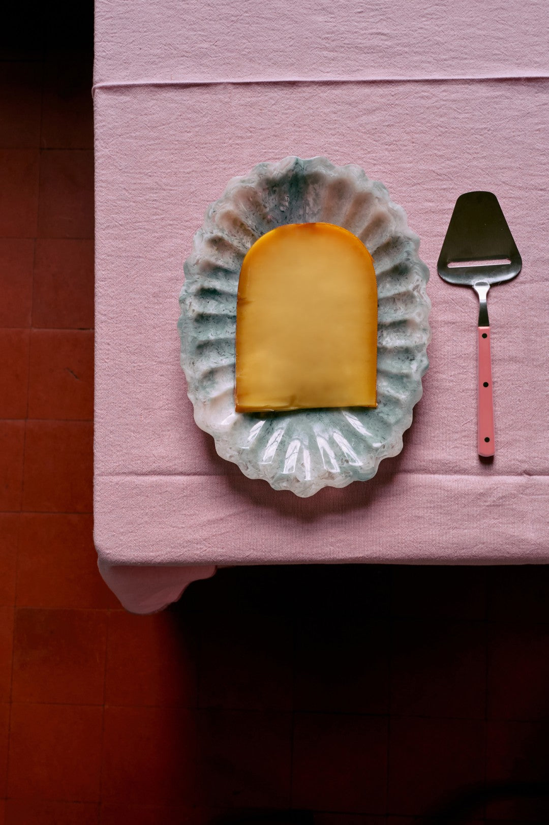Rectangular piece of cheese on a decorative plate with a pink spatula on a pink surface.