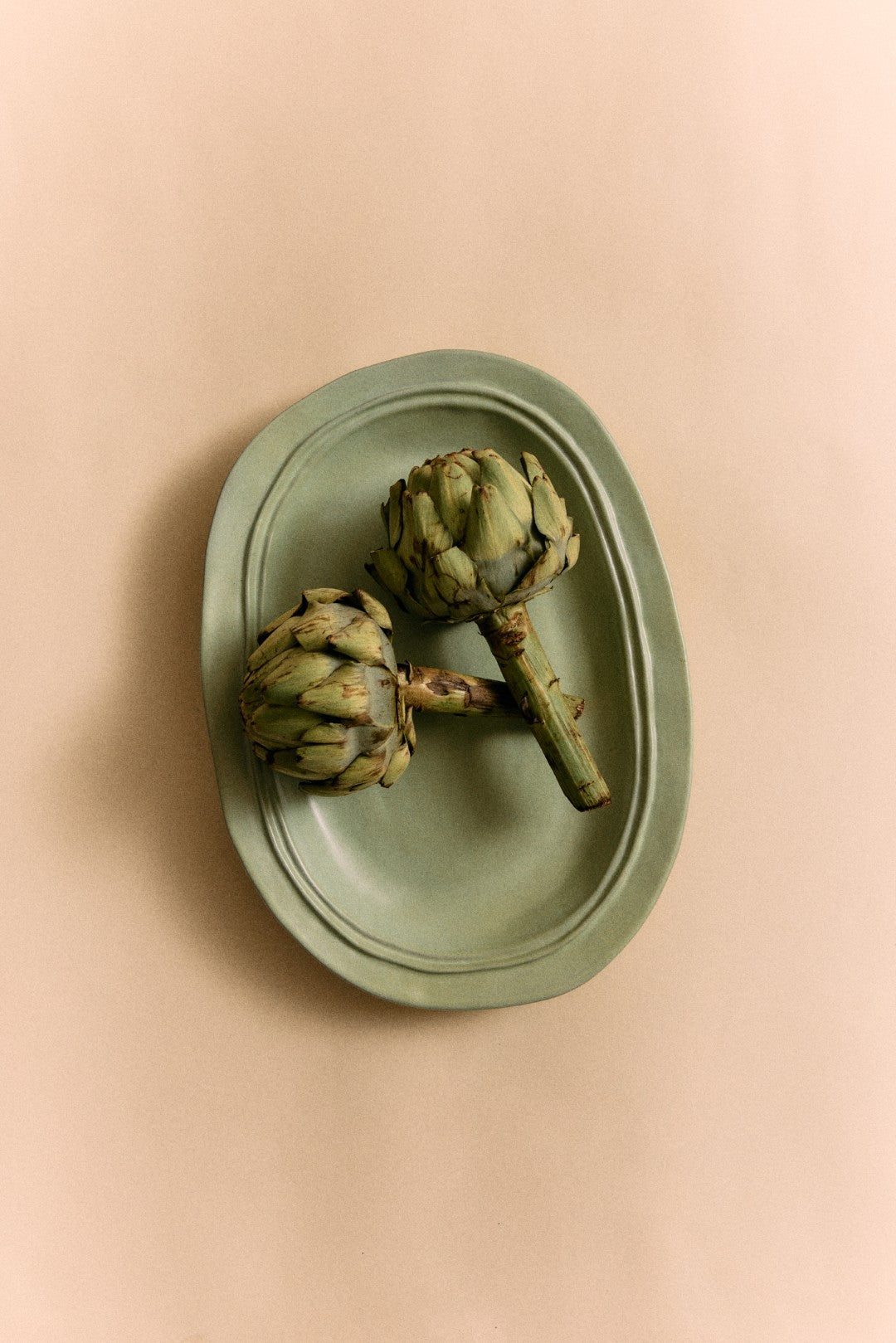 Two artichokes on a green plate against a beige background