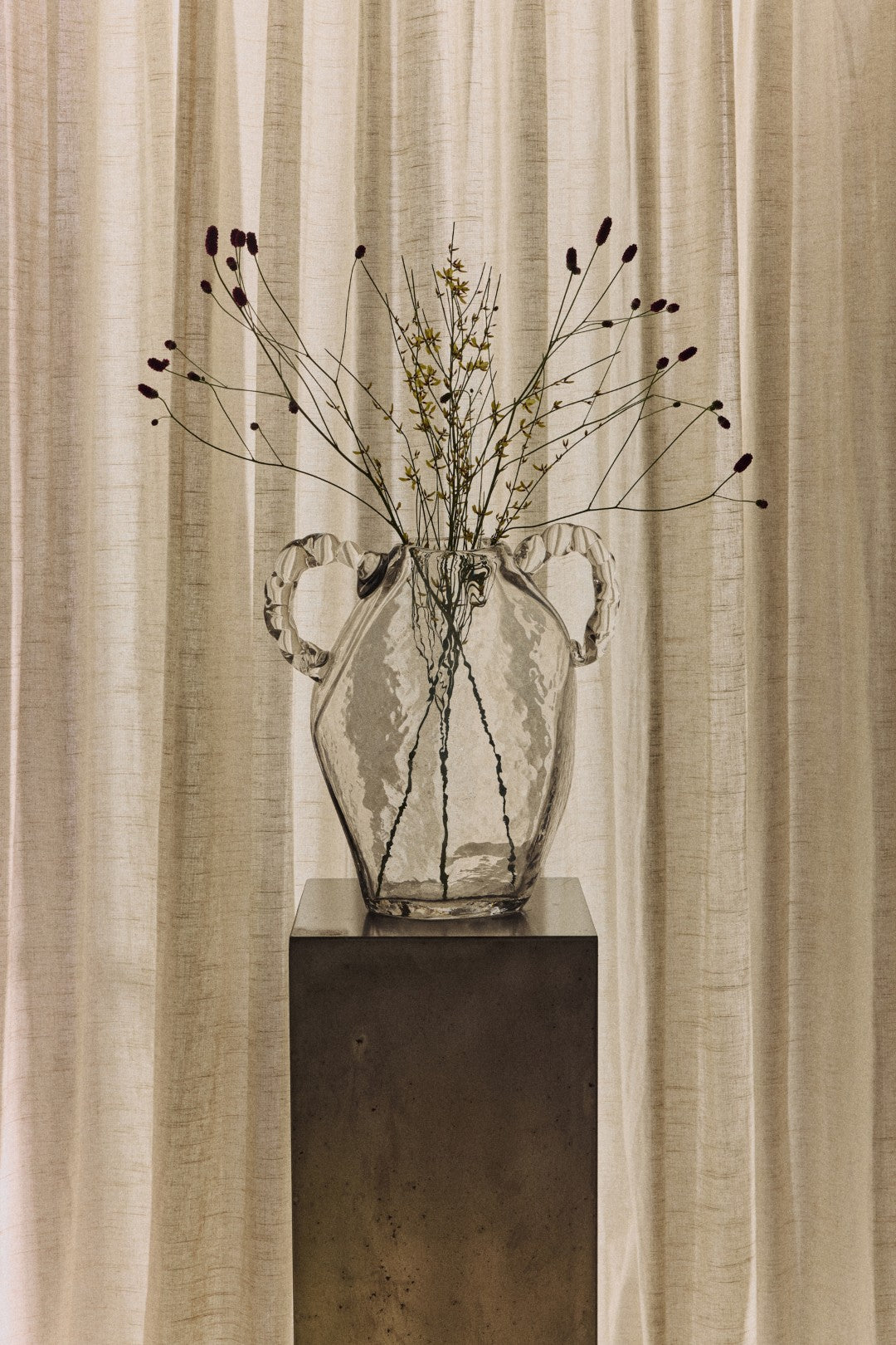A clear glass vase with two handles sits on a dark pedestal in front of draped beige curtains, holding dried stems and small buds.