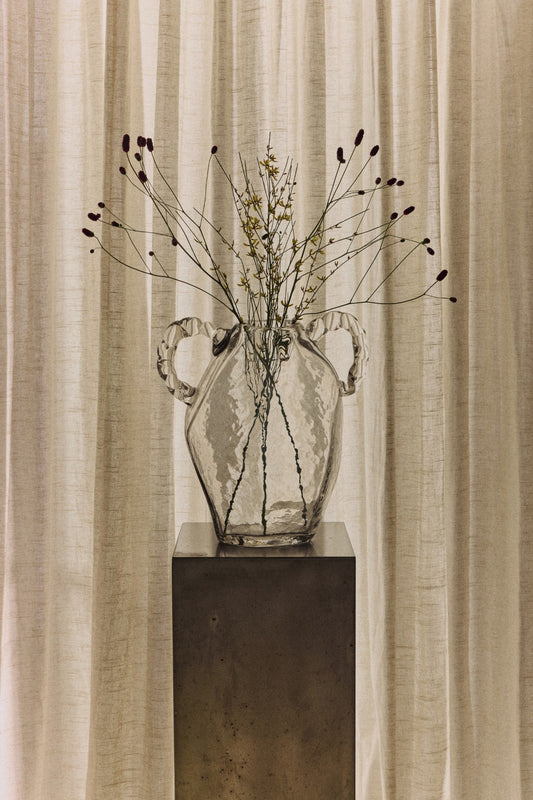 A clear glass vase with two handles sits on a dark pedestal in front of draped beige curtains, holding dried stems and small buds.
