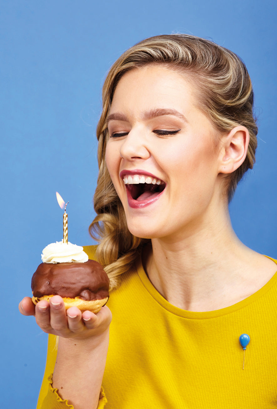 woman holding a piece of cake with a candle wearing a blue balloon pin on yellow sweater