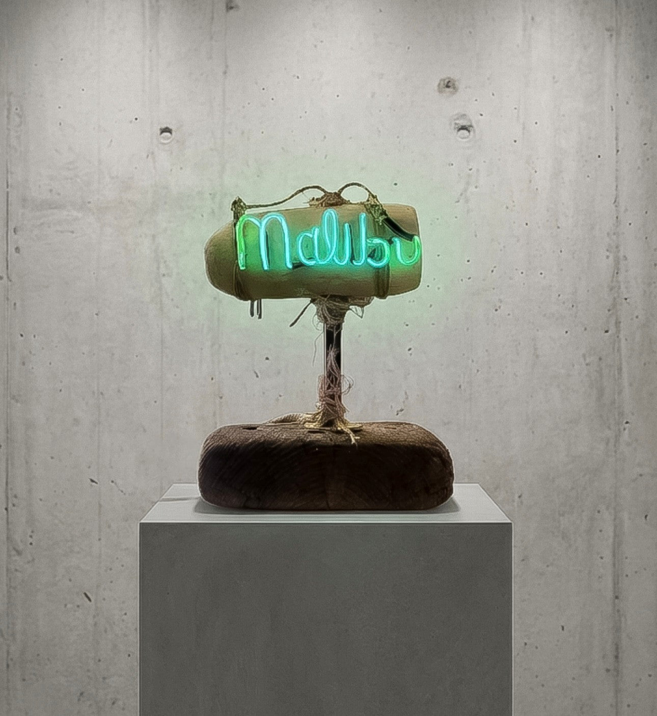 A sculptural neon sign reading 'Malibu' in bright green, mounted on a rustic wooden base with a textured, earthy finish, displayed on a white pedestal against a concrete wall.