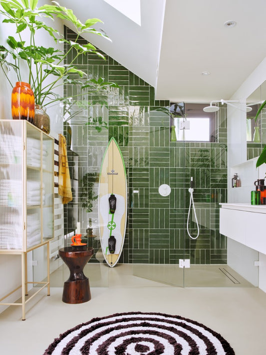 bathroom with green tile in shower, surfboard, burgundy wooden stool and black and white round bathmat