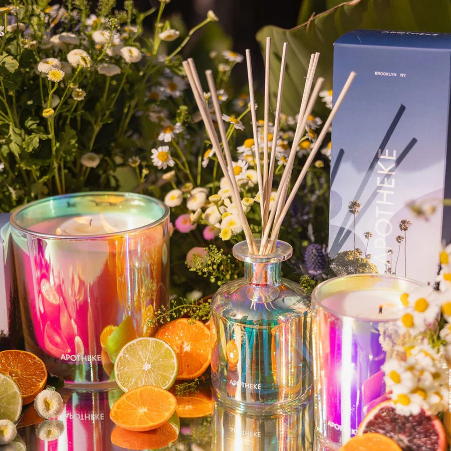 Aromatherapy diffuser with reeds, colorful candles, and fruits on a reflective surface with a blurred floral background.