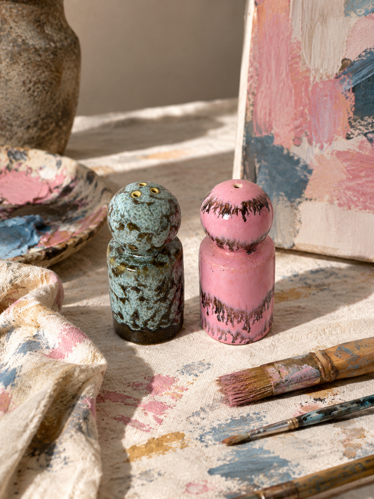 Two small hand-painted ceramic figurines, one in textured teal with gold accents and one in pink with dark brown drips, displayed on a paint-splattered cloth with paintbrushes and an artist's palette in the background.