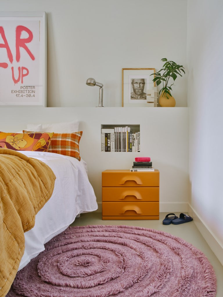 round lilac rug with high and low piles nect to a bed and an nightstand with 3 orange drawers