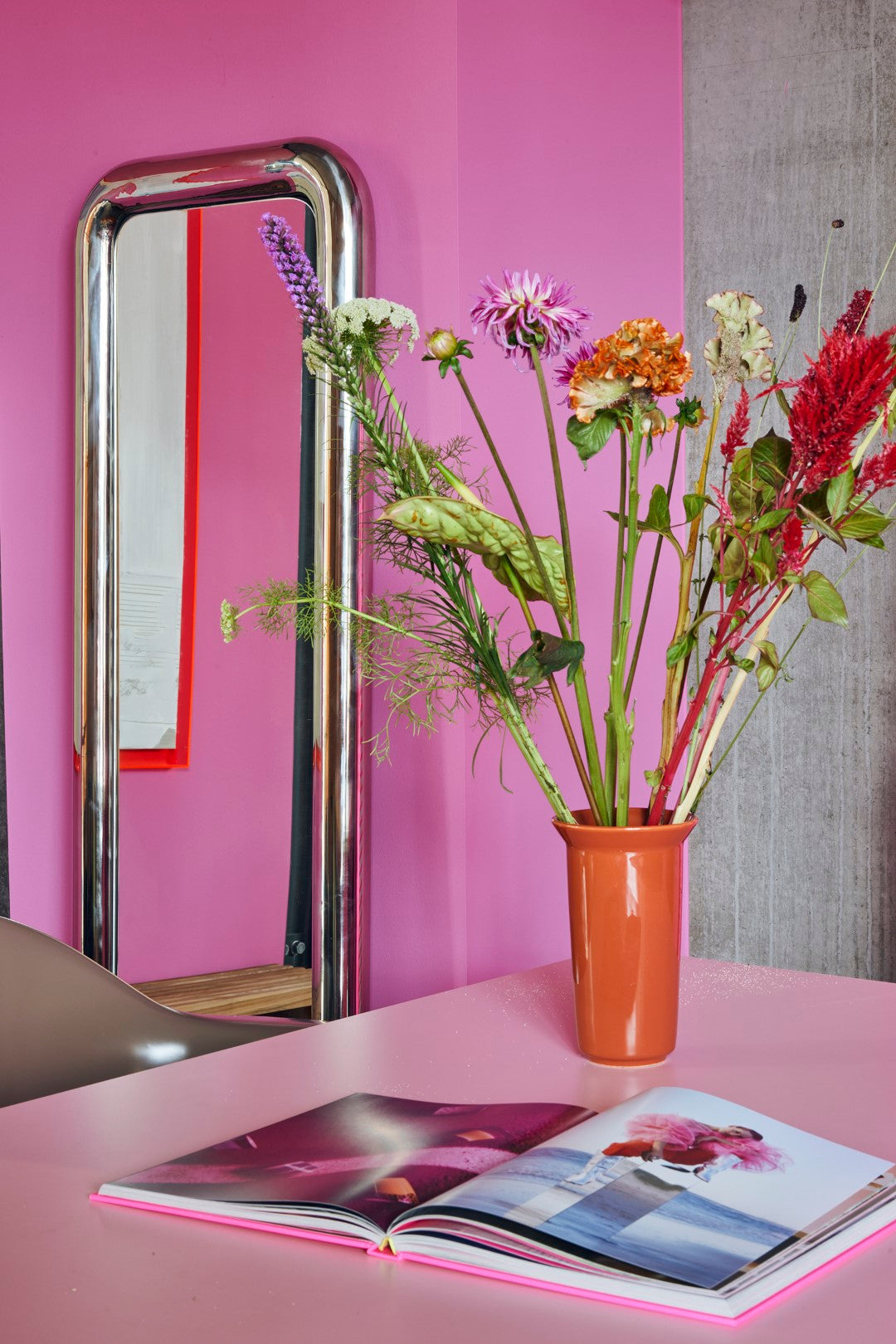 tall long standing chrome chubby mirror on pink wall and orange vase with flowers