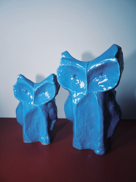 Two blue ceramic owl figurines on a dark surface with a light gray background