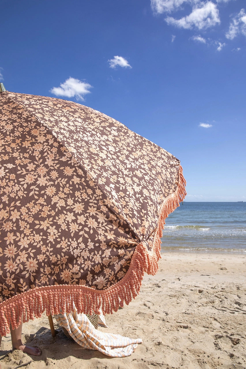 retro style beach umbrella with brown and orange floral fabric and orange fringes at the beach with a blue sky with a few small clouds
