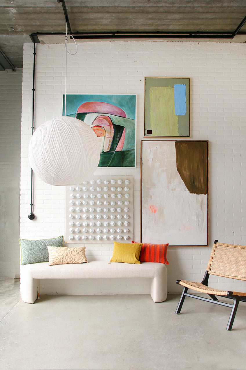 green printed pillow with yellow trim piping on a white linen bench underneath a gallery wall with abstract paintings